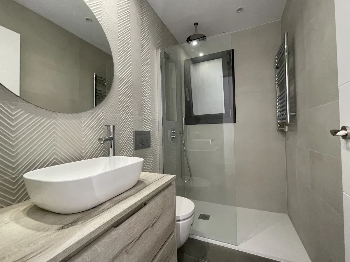 Segundo baño reformado con amplio plato de ducha, mueble lavabo acabado madera y revestimiento con patrón zigzag.