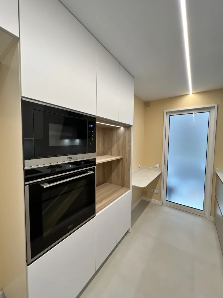 Cocina vista horno y mesita: Columna con horno y microondas integrados, estantería de madera y mesita de desayuno en esta reforma completa de vivienda.