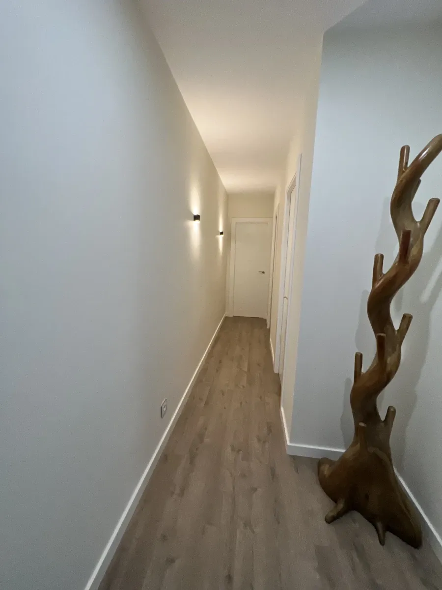 Pasillo luminoso con apliques de pared modernos, suelo laminado y zócalos blancos, resultado de una reforma completa de vivienda.